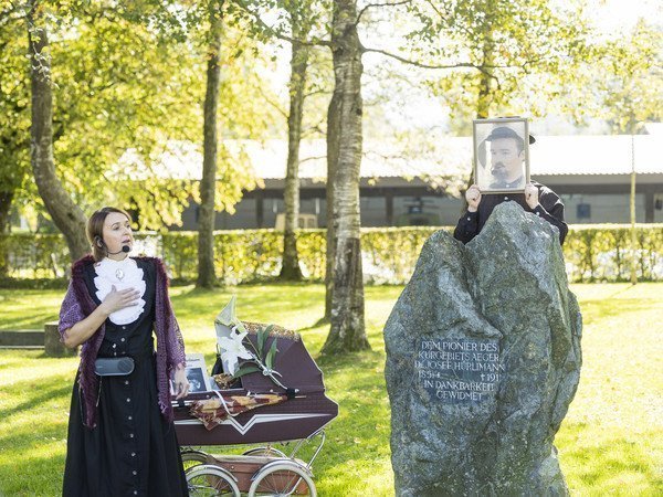 Frau in altem, edlem Kleid mit Kinderwagen in Park vor einem Stein